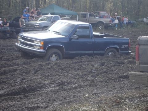 1989 Ford Bronco - A blue pickup truck is stuck deep in the mud, with spectators watching from the side and other vehicles in the background.