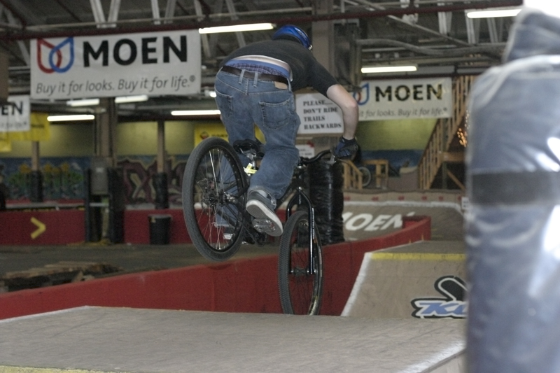 Person on a bicycle performing a trick on a ramp indoors. Signs for MOEN are visible