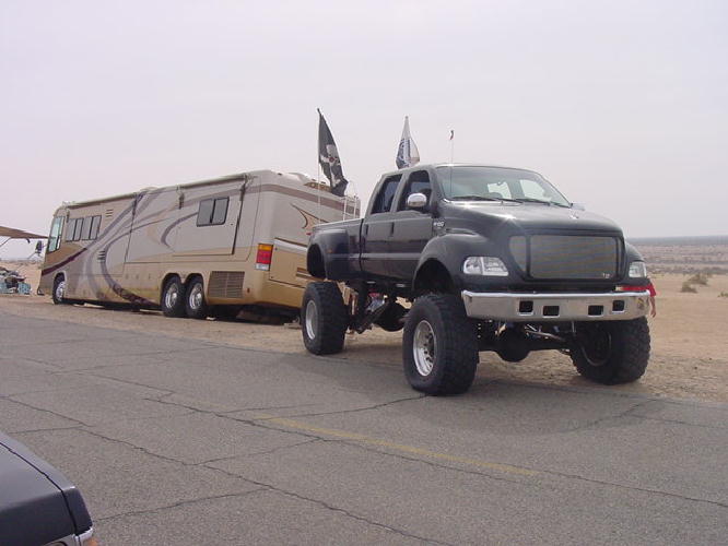 Lifted black Ford crew cab pickup on big tires parked beside a tan motorhome in the desert.