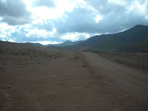 1988 Ford Bronco - A desolate dirt road winds through a dry, hilly terrain with distant mountains under a dramatic, cloudy sky.