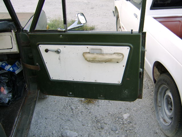 Green vehicle door interior with white panel, chrome handle, and worn armrest. Another white truck is visible to the right