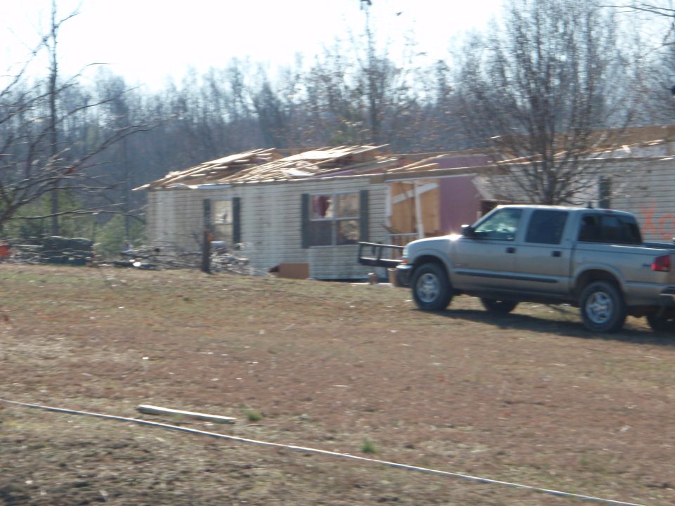 Silver crew-cab pickup parked beside a storm-damaged house with roof torn off