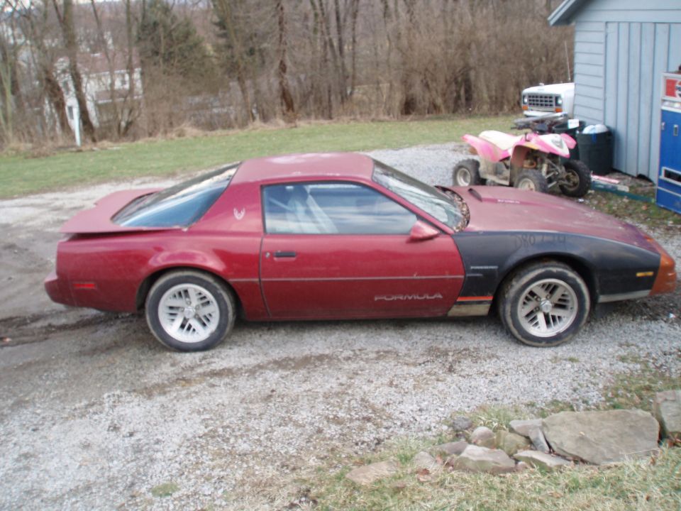 Red and black 1991 Pontiac Firebird Formula with a spoiler and aftermarket wheels, parked on a gravel driveway