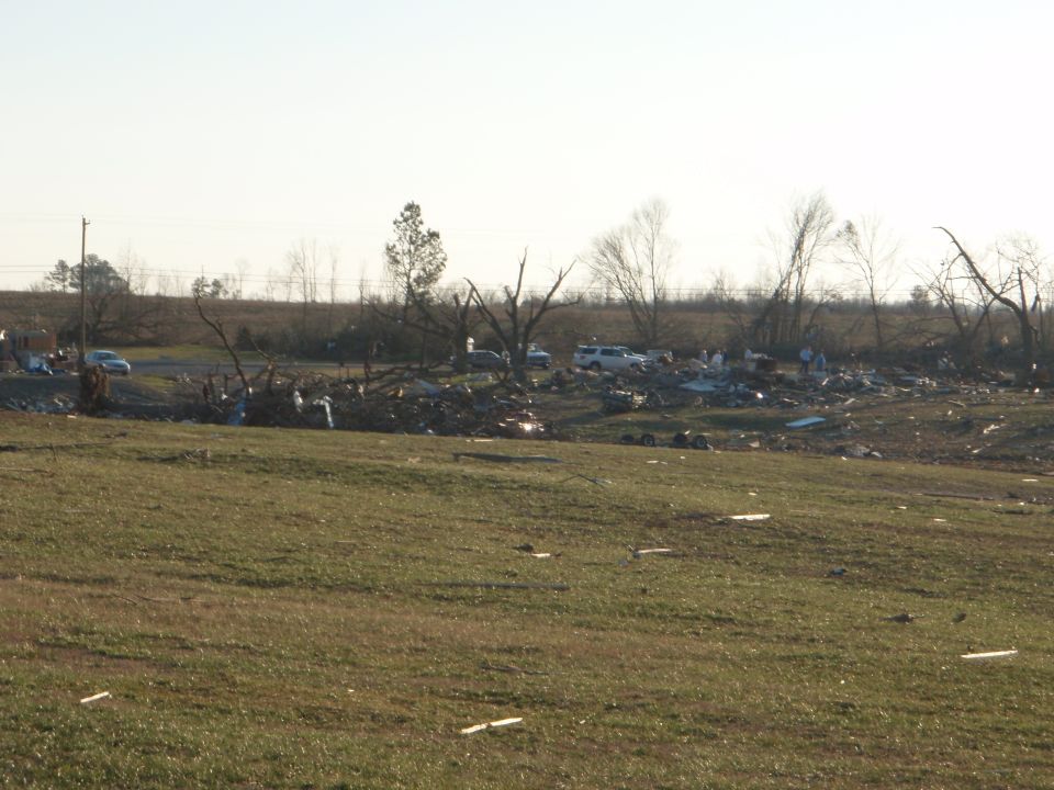 Storm-damaged field with debris, broken trees, and distant parked vehicles