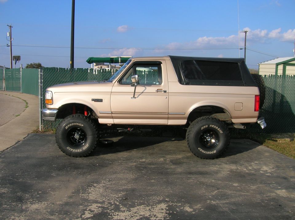 Tan lifted two-door Ford Bronco, left-side profile on black mud tires