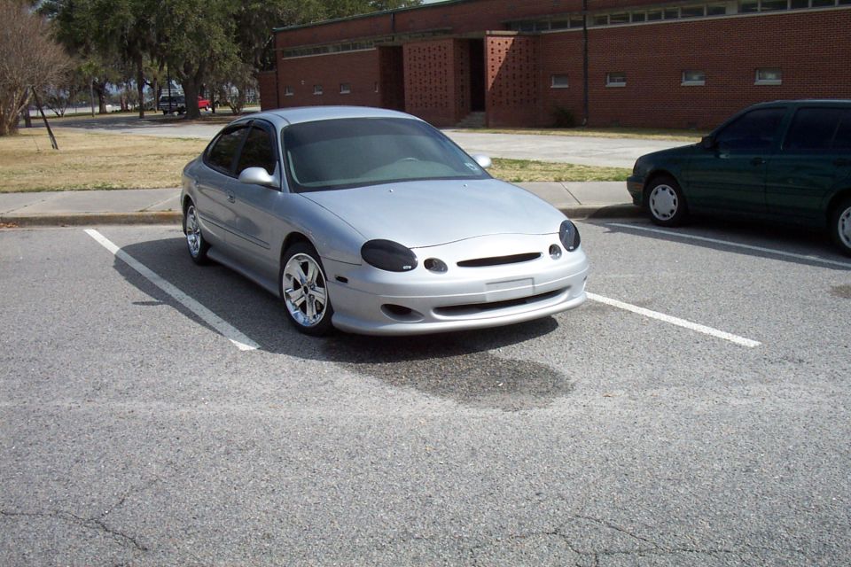 Silver sedan parked in a lot, front end visible. Another dark green vehicle is parked to its right