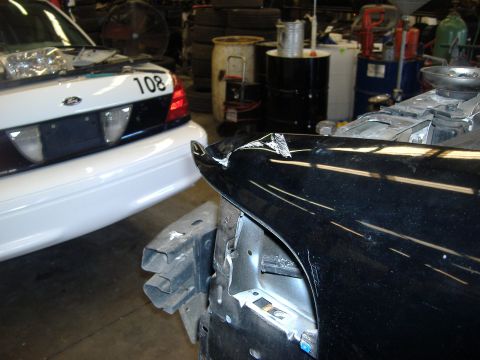 2001 Ford Crown Victoria - Two Ford Crown Victorias are in a repair shop, one with a damaged black fender and the other white with a visible number.