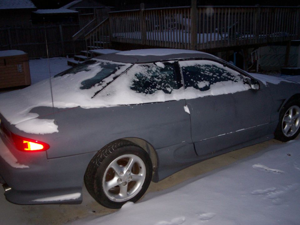 Side view of a gray coupe with snow on the roof and windows, parked outside in winter