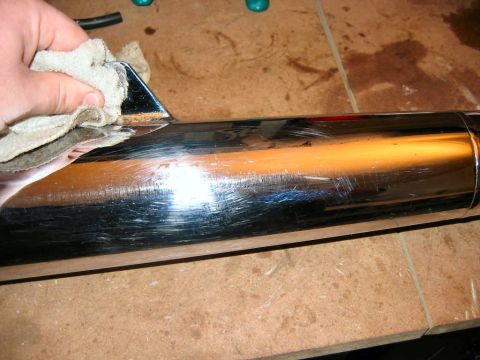 1985 Ford LTD Crown Victoria - A hand wipes a heavily scratched chrome exhaust pipe, revealing rust on its bracket.
