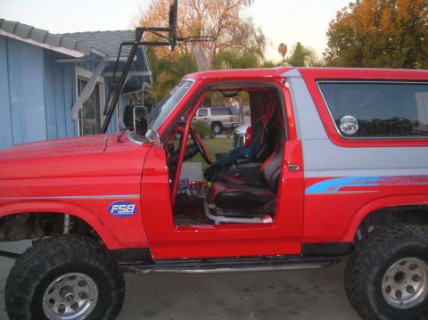 1985 Ford Bronco - This red SUV has its driver's door open, revealing racing seats and a roll cage. Off-road tires are visible on the front and rear.