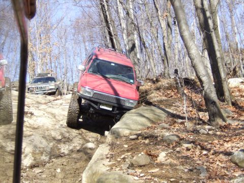 1976 Toyota Land Cruiser - Two SUVs tackle a challenging off-road course through a dense forest on a sunny day.