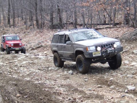 1976 Toyota Land Cruiser - Two Jeep SUVs navigate a challenging rocky and muddy off-road trail through a wooded area.