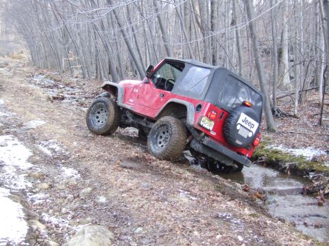 1976 Toyota Land Cruiser - A red Jeep Wrangler navigates a challenging off-road trail covered in mud and scattered snow.