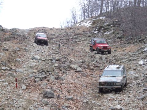 1976 Toyota Land Cruiser - Several SUVs navigate a challenging, boulder-strewn path on a wooded hillside.