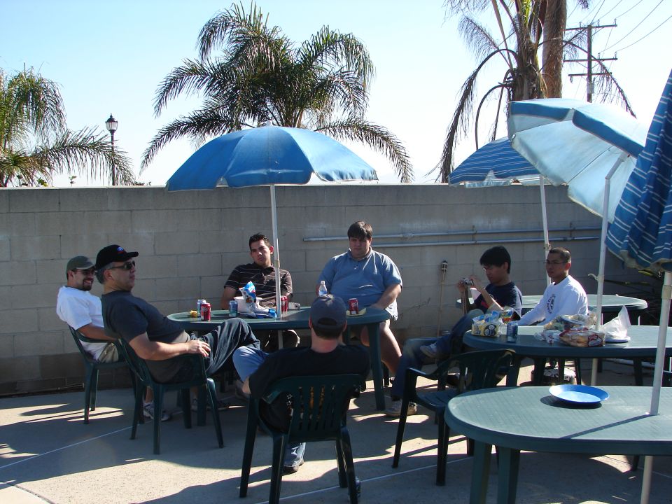 People at green patio tables under blue umbrellas in a walled backyard