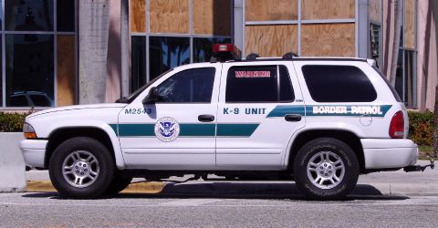 1991 Ford Crown Victoria - A white SUV marked "Border Patrol K-9 UNIT" is parked in front of a building with boarded-up windows.