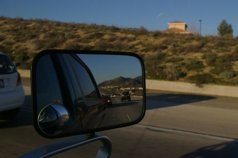 1986 Ford Bronco - Closeup of a rectangular black tow-mirror with a stick-on round convex spot mirror, shot from inside a moving vehicle on a freeway. The mir…