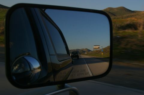 1986 Ford Bronco - A truck is seen in the side mirror as it travels down a highway with hills in the distance.