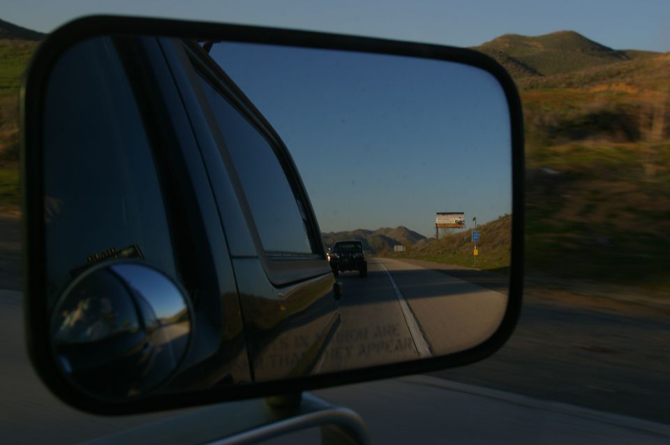 view from side mirror of truck on highway, another truck ahead, hills in background