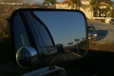 1986 Ford Bronco - Close-up of a black towing-style side mirror with a round convex spot mirror, reflecting the vehicle's own body line and a dark pickup foll…