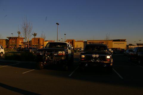 1986 Ford Bronco - Two trucks are parked in a lot with a commercial building in the background.