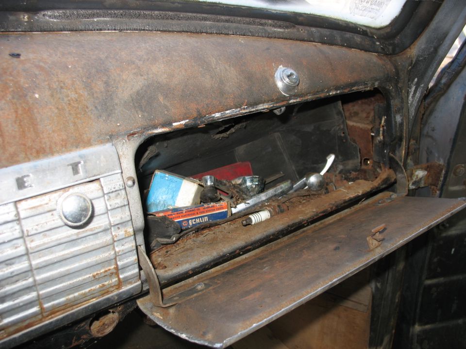 Open glove box filled with old parts, including spark plugs and boxes. The dashboard is rusty