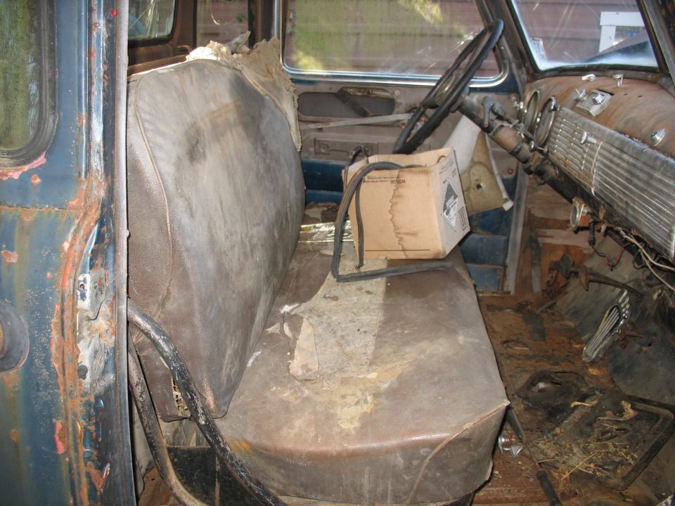 Interior of a worn-out truck cab, showing a bench seat, steering wheel, and dashboard. The interior is rusty and shows signs of disrepair