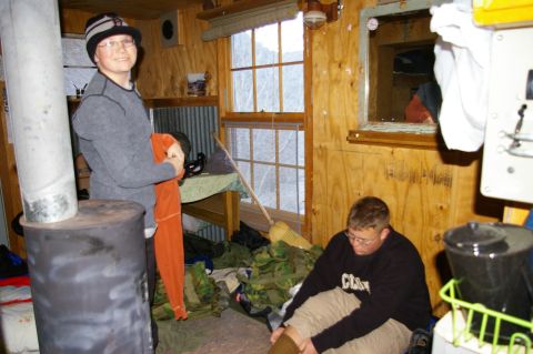 1986 Ford Bronco - Two people are inside a rustic cabin, one standing near a wood stove and the other sitting on the floor.