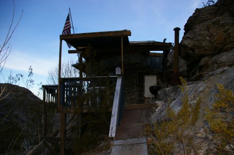 1986 Ford Bronco - A rustic cabin is perched on a steep, rocky slope under a clear blue sky, with an American flag waving from its roof.