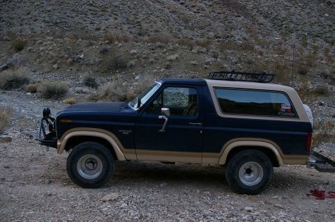 1986 Ford Bronco - A dark blue and tan Ford Bronco is parked on a gravelly surface with a rocky hillside in the background.
