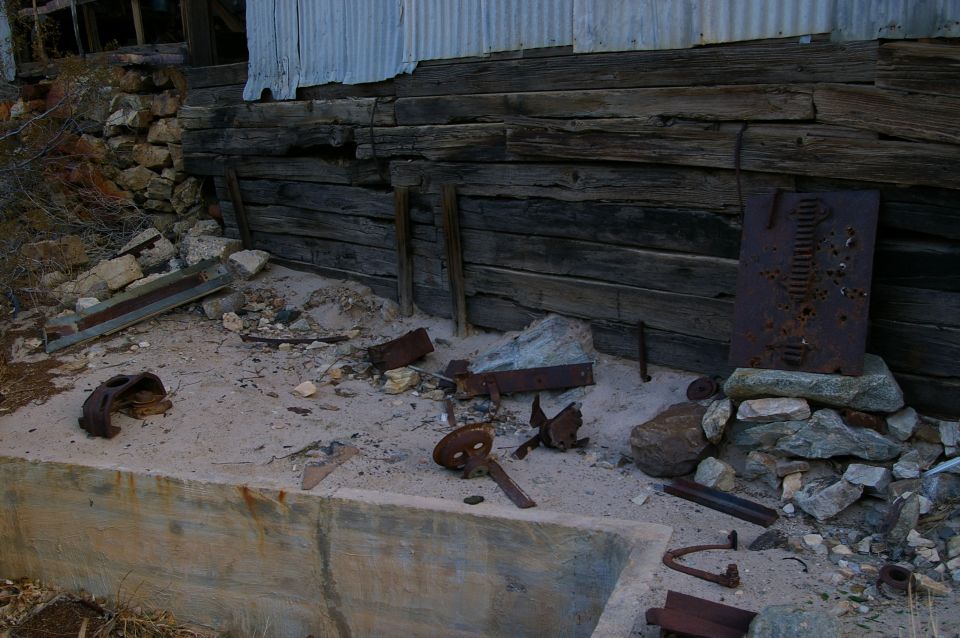 Debris and rusted metal parts scattered on sand in front of a weathered wooden wall and corrugated metal structure