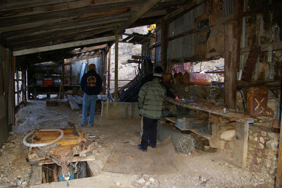 Dilapidated interior of a workshop with two people, rusty barrels, and scattered tools