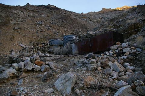 1986 Ford Bronco - Two corrugated metal water tanks and a large rusted rectangular tank sit amidst rocks and sparse vegetation in a desert environment.