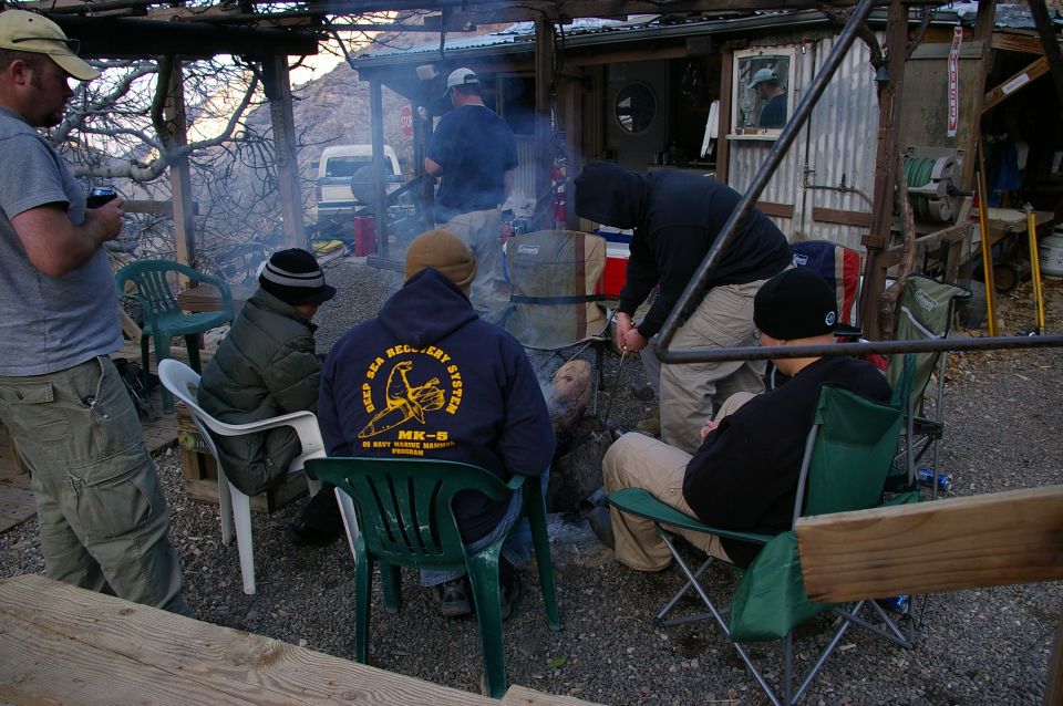 People sit around a fire pit outside a rustic structure. One person wears a hoodie with "Deep Sea Recovery System MK-8" on it