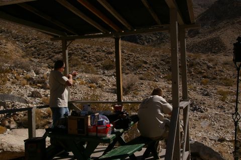 1986 Ford Bronco - Men are at a covered picnic table in a desert landscape, with one man aiming a rifle.