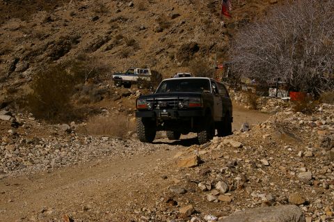 1986 Ford Bronco - Two off-road vehicles navigate a dusty, rocky trail in a desert environment under a clear sky.