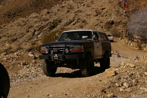 1986 Ford Bronco - A two-tone Ford Bronco drives on a rocky desert road, with another vehicle visible in the distance.