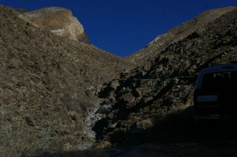 1986 Ford Bronco - A dark-colored SUV is parked on a dirt road, with steep, rocky canyon walls rising on either side under a clear blue sky.