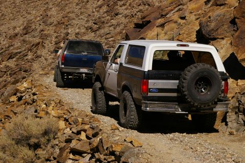 1986 Ford Bronco - A Ford Bronco and another SUV navigate a rough, rocky trail in a desert landscape.
