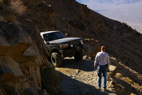 1986 Ford Bronco - A man observes a Ford F-Series truck navigating a steep, rocky mountain trail with a vast desert landscape in the background.