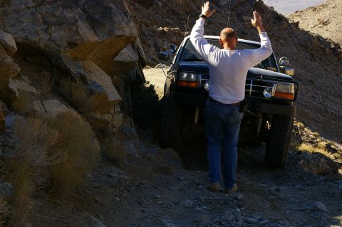 1986 Ford Bronco - A man stands with his arms up next to a black SUV navigating a rough, rocky terrain.