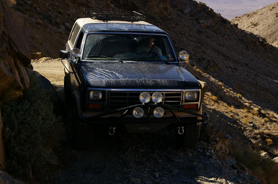 Ford Bronco driving on a dirt road in a canyon. It has a roof rack and extra lights on the front