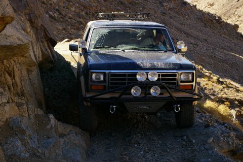 1986 Ford Bronco - A dark blue Ford Bronco navigates a narrow, rocky path in a desert landscape.