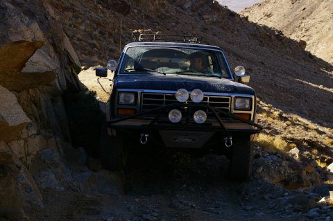 1986 Ford Bronco - This 1986 Ford Bronco navigates a narrow, rocky mountain path, its front grille and auxiliary lights clearly visible.