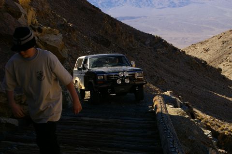 1986 Ford Bronco - A dark-colored Ford Bronco navigates a rough mountain road, with a person walking ahead on a wooden path.