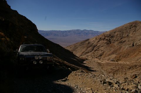 1986 Ford Bronco - A dark SUV is parked on a rough dirt road in a mountainous desert landscape under a clear blue sky.