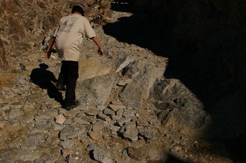 1986 Ford Bronco - A person walks along a , rocky trail under bright sunlight, casting a long shadow.