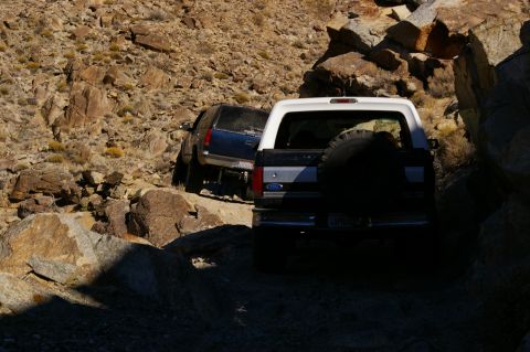 1986 Ford Bronco - Two SUVs navigate a narrow, rocky trail in a desert landscape.