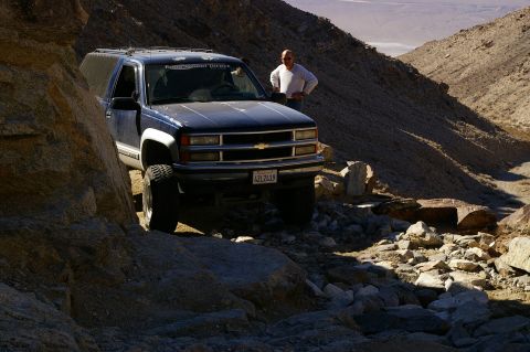 1986 Ford Bronco - Man stands by a dark blue Chevrolet SUV navigating a rocky desert trail. The vehicle appears to be from the mid-1990s.