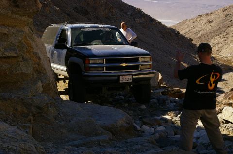 1986 Ford Bronco - A man waves from the side of a rocky trail as a Chevrolet Suburban navigates the difficult terrain.